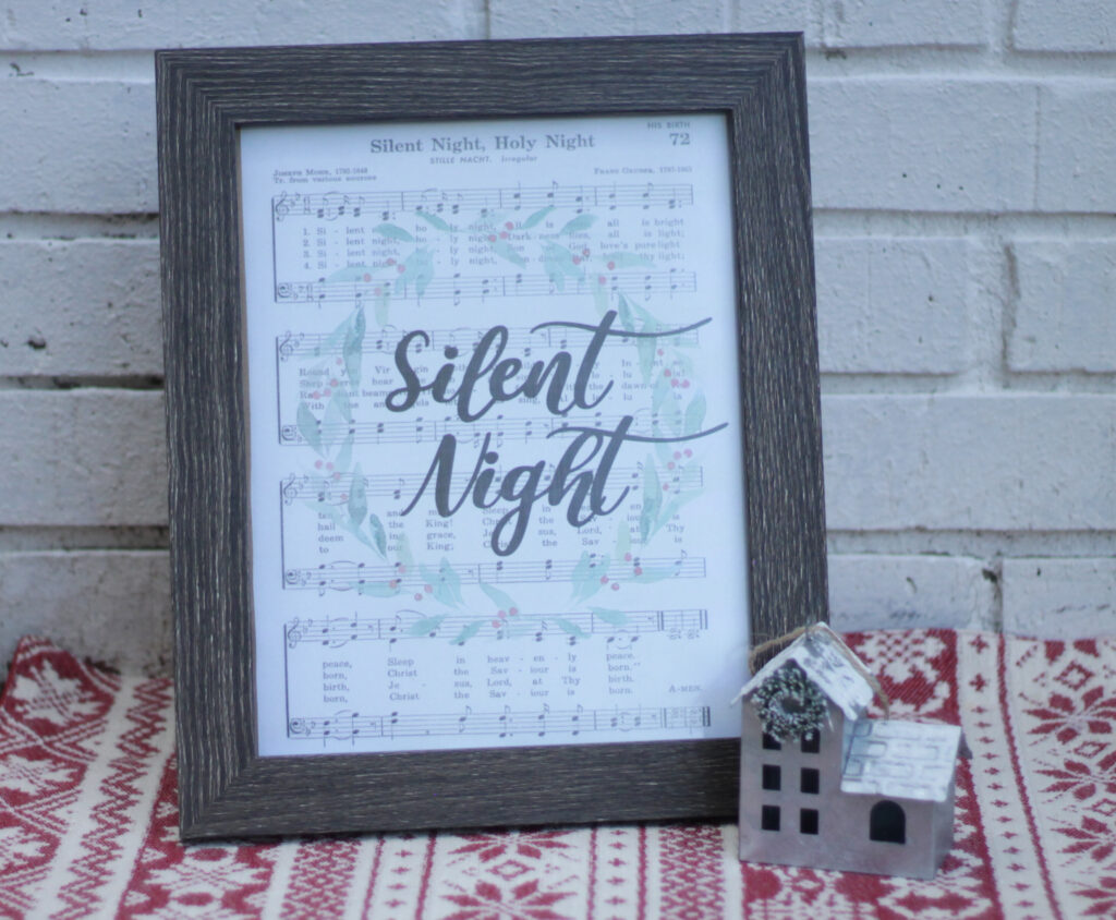 wooden frame with Silent night sheet music inside displaying a Christmas wreath and script text of silent night in the center, beside a metal house decor on top of red and cream Christmas designed fabric 