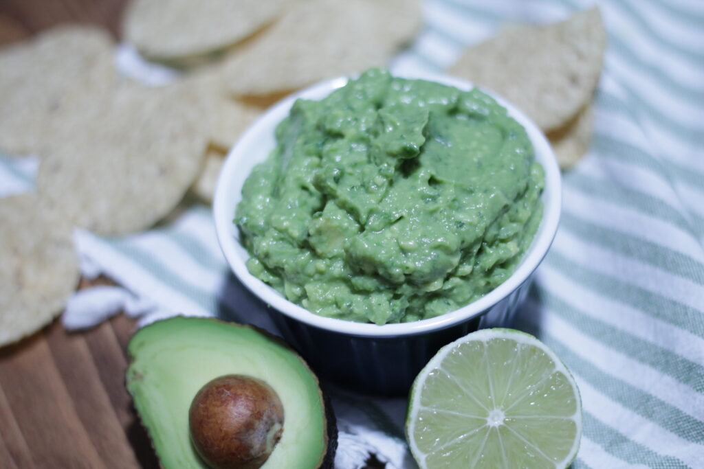 Guacamole in a small blue bowl, half of an avocado and half of a lime sitting in front of it, with tortilla chips scattered behind the bowl.