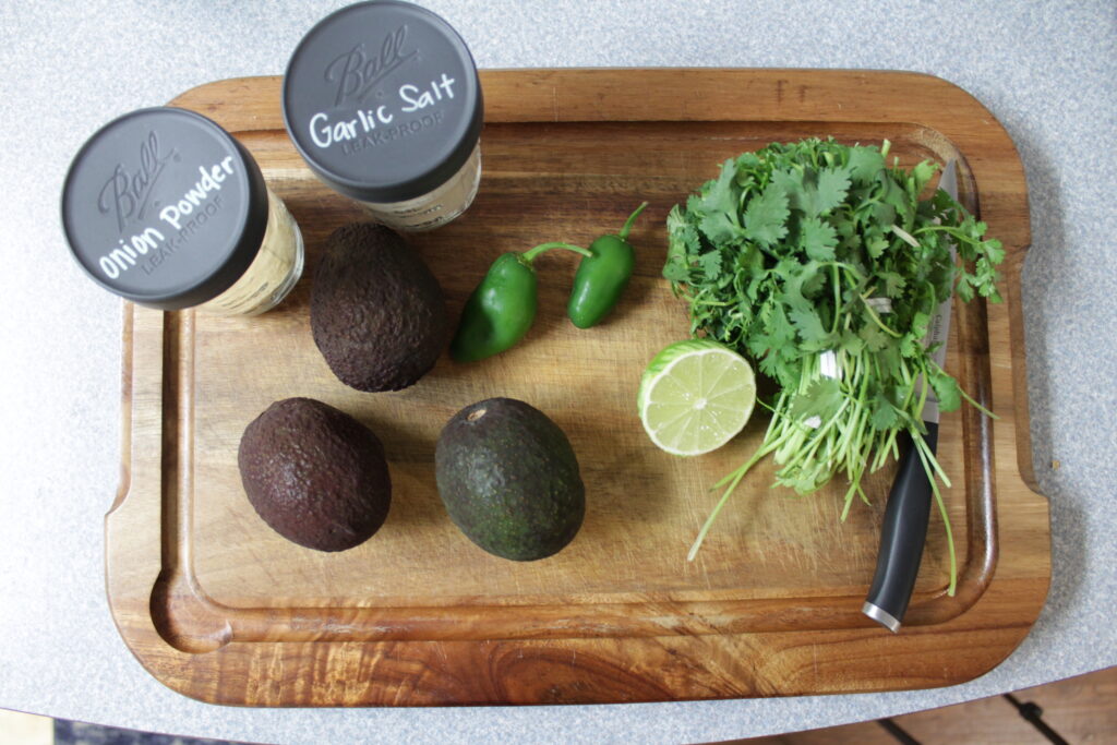 Onion powder, garlic salt, 3 avocados, 2 small jalapeños, half of a lime, one bunch of cilantro, and a knife sitting on wooden cutting board