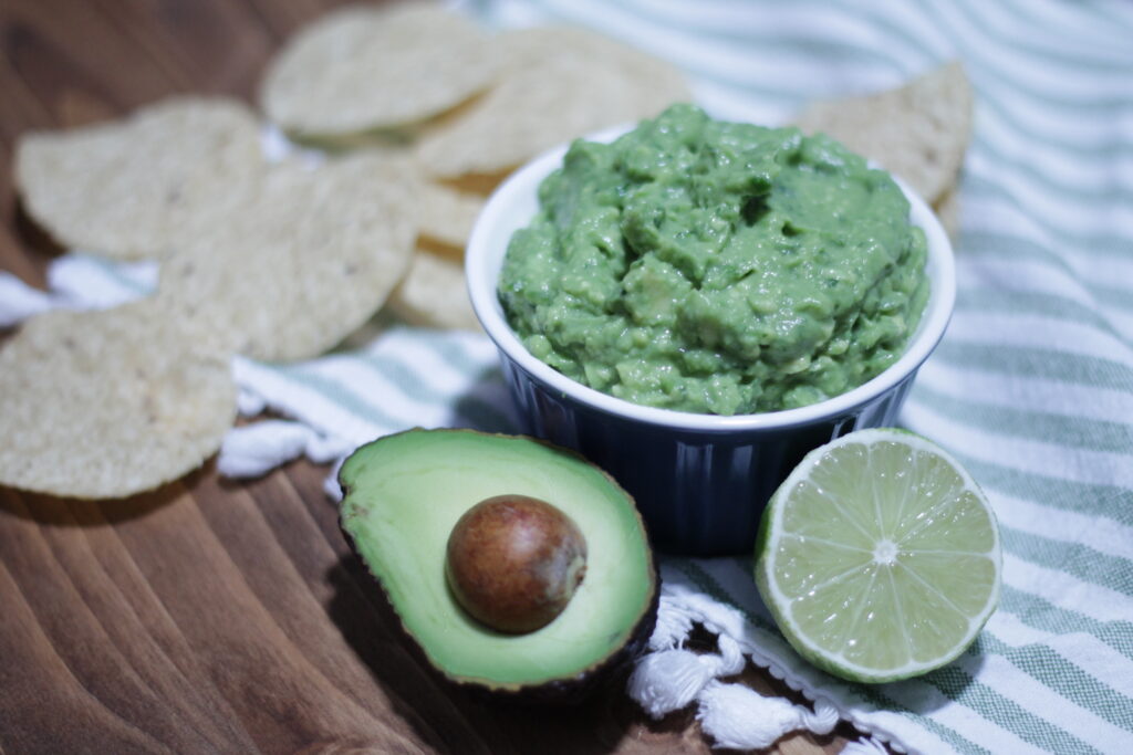 Guacamole in a small blue bowl, half of an avocado and half of a lime sitting in front of it, with tortilla chips scattered behind the bowl.