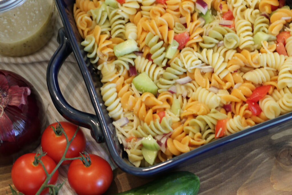 pasta salad in blue dish with onion, tomatoes, and cucumber on the left.