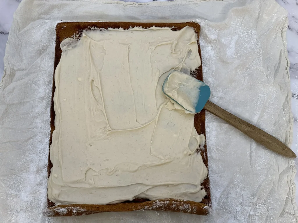 spreading cream cheese icing over pumpkin roll cake 